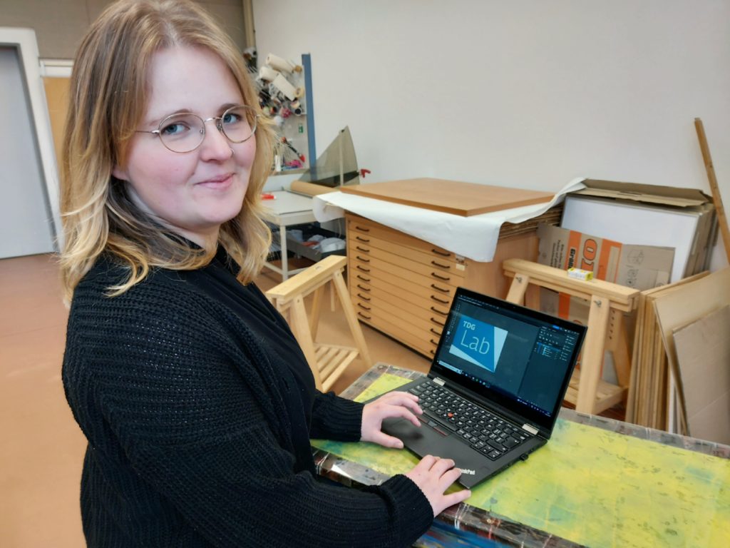 Studentin Julia Seidler zeigt auf einem Laptop das neue Logo des TDG-Labs, das sie designt hat.